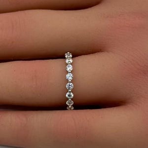 May include: A silver-toned eternity band ring featuring a continuous row of small, round, clear faceted stones set in a metallic channel. The ring is thin and delicate, showcasing a uniform design with sparkling stones that wrap around the band.