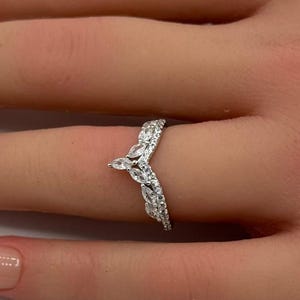 May include: A silver-toned V-shaped ring featuring a series of marquise-cut and round-cut clear stones arranged in a pointed chevron design. The band is slender and metallic, showcasing a symmetrical arrangement of sparkling stones along the top.
