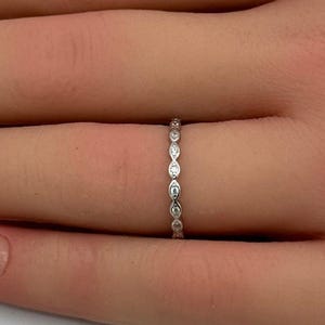May include: A thin silver-toned band ring featuring a series of small, marquise-shaped settings, each containing a tiny, clear, sparkling stone. The ring is worn on a finger, showcasing the delicate, repeating geometric pattern of the metalwork.