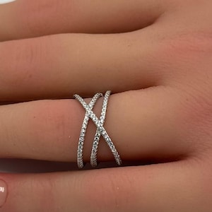 May include: A silver-toned crossover ring featuring two intersecting bands encrusted with small, clear, sparkling stones. The design creates an X-shaped pattern across the finger, with the metallic finish providing a bright, reflective surface.
