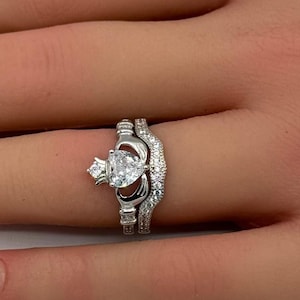 May include: A silver-toned Claddagh ring featuring a central heart-shaped clear gemstone, a crown detail, and two hands. The band is split into two parts, with the outer band adorned with small, clear pavé-set stones along the entire visible surface.
