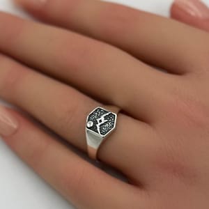 Puede incluir: Un anillo de sello plateado con una cara hexagonal. La cara presenta un patrón oscuro y texturizado y un pequeño acento brillante. El anillo se lleva en un dedo, con un fondo suave de flores rosas.