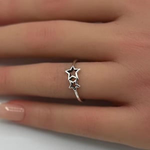 May include: A silver ring featuring two star-shaped charms. The stars have a black interior and are connected to the ring band. The ring is shown on a hand with manicured nails, against a white background with pink flowers.