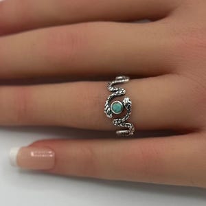 Peut inclure: Bague argentée ornée d'un motif de serpent, avec une petite pierre turquoise ronde au centre. La bague est portée sur une main aux ongles manucurés, sur fond blanc avec des fleurs roses.