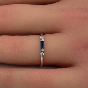 May include: A thin silver-toned ring featuring a central rectangular baguette-cut dark blue gemstone flanked by two small clear round stones on each side, set in a minimalist metal band that rests on a finger.