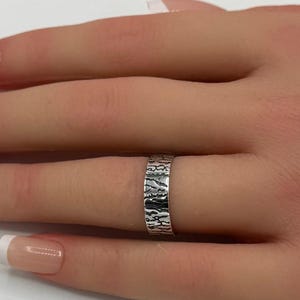 May include: A silver ring with a textured design is worn on a hand. The ring has a band-style design with a detailed, possibly etched, pattern. The hand has manicured nails with a French tip design. Pink flowers are in the background.