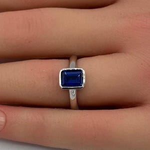 May include: A silver-toned ring featuring a rectangular, emerald-cut blue gemstone set in a smooth bezel setting. The band is plain and metallic, showcasing a clean, polished finish that encircles the finger with a simple, centered design.