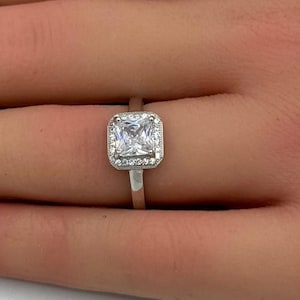 May include: A silver-toned ring featuring a square-cut clear center stone surrounded by a halo of small, round, clear accent stones. The band is thin and metallic, providing a clean, polished look that highlights the central square setting.