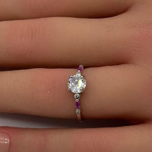 May include: A silver-toned ring featuring a central round clear stone, flanked by smaller clear stones and vibrant pink accent stones set along the band. The ring is displayed on a finger, showcasing the metallic band and the arrangement of the stones.
