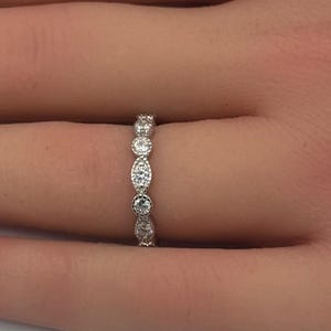 May include: A silver-toned band ring featuring an alternating pattern of round and marquise-shaped clear stones. The metal setting displays a delicate milgrain texture along the edges, creating a refined and detailed appearance on the finger.