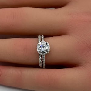 May include: A silver-toned engagement ring set featuring a round brilliant-cut center stone surrounded by a halo of small pavé-set stones. The band and the matching wedding band are both encrusted with tiny, sparkling pavé-set stones throughout.