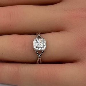 May include: A silver-toned ring featuring a central square-cut stone surrounded by a halo of smaller round stones. The band has a split, crossover design that tapers toward the center setting, creating a sleek and polished appearance on the finger.