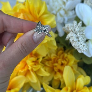 Op de afbeelding: Een zilveren vlinderring met gedetailleerde vleugelpatronen. De ring wordt getoond tegen een achtergrond van gele en witte bloemen. Het vlinderontwerp is het middelpunt van de ring.