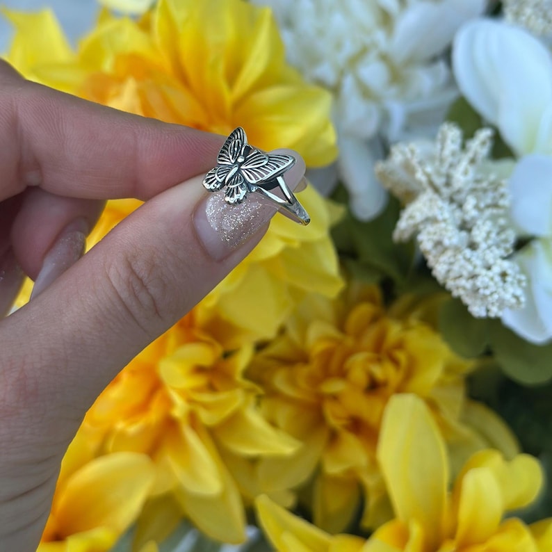 Op de afbeelding: Een zilveren vlinderring met gedetailleerde vleugelpatronen. De ring wordt getoond tegen een achtergrond van gele en witte bloemen. Het vlinderontwerp is het middelpunt van de ring.