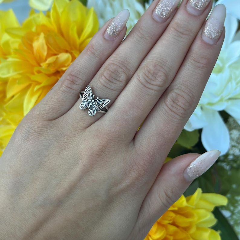 Op de afbeelding: Een zilveren vlinderring. De ring heeft een gedetailleerd vlinderontwerp met open vleugels. De ring wordt aan een vinger gedragen, met een achtergrond van gele en witte bloemen.
