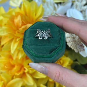 Op de afbeelding: Een zilveren vlinderring gepresenteerd op een groene fluwelen ringdoos. De ring heeft een gedetailleerd vlinderontwerp met open vleugels. De achtergrond bevat gele en witte bloemen, wat een levendig contrast cre&euml;ert.