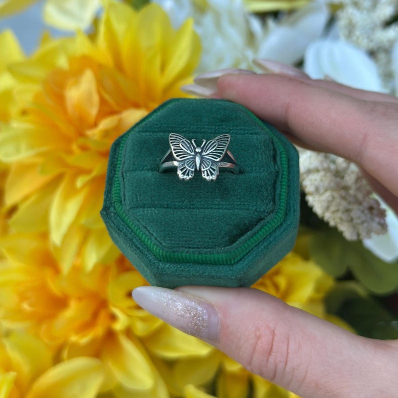Op de afbeelding: Een zilveren vlinderring gepresenteerd op een groene fluwelen ringdoos. De ring heeft een gedetailleerd vlinderontwerp met open vleugels. De achtergrond bevat gele en witte bloemen, wat een levendig contrast cre&euml;ert.