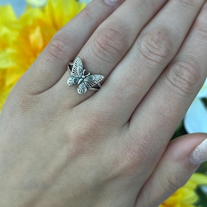 Op de afbeelding: Een zilveren vlinderring. De ring heeft een gedetailleerd vlindermotief met opengewerkte vleugels en een getextureerd lichaam. De ring wordt aan een vinger gedragen en toont de ingewikkelde details van het sieraad.