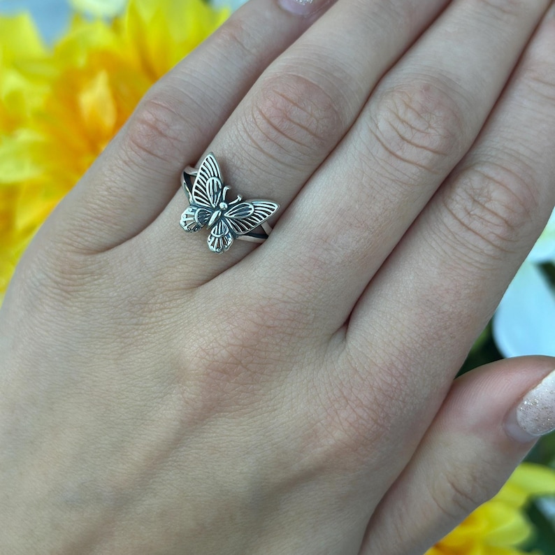 Op de afbeelding: Een zilveren vlinderring. De ring heeft een gedetailleerd vlindermotief met opengewerkte vleugels en een getextureerd lichaam. De ring wordt aan een vinger gedragen en toont de ingewikkelde details van het sieraad.