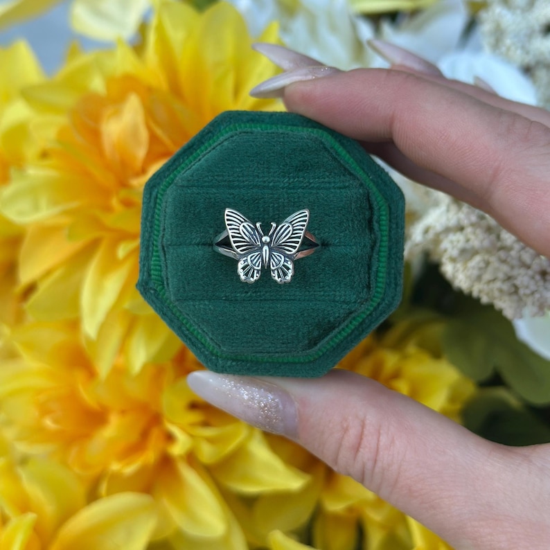 Op de afbeelding: Een zilveren vlinderring gepresenteerd in een groene fluwelen ringdoos. De ring heeft een gedetailleerd vlinderontwerp. De achtergrond bestaat uit gele en witte bloemen.