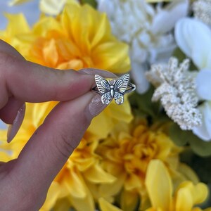 Op de afbeelding: Een zilveren vlinderring met gedetailleerde vleugelpatronen. De ring wordt vastgehouden voor een wazige achtergrond van gele en witte bloemen. Het vlinderontwerp is het middelpunt en toont ingewikkelde lijnen en rondingen.
