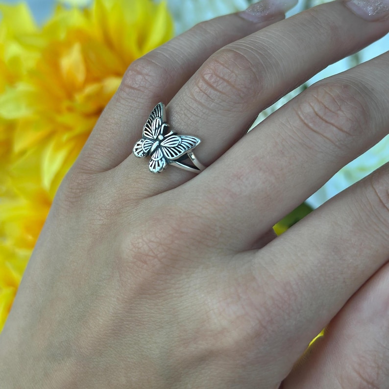 Op de afbeelding: Een zilveren vlinderring met gedetailleerde vleugelpatronen. De ring wordt aan een hand gedragen, waarbij het vlinderontwerp prominent aanwezig is. De band van de ring is dun en eenvoudig, als aanvulling op de ingewikkelde vlinder. De achtergrond heeft gele bloemen.