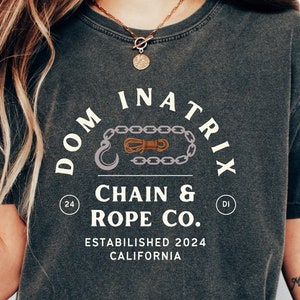 May include: A dark grey t-shirt with a white graphic design featuring a chain and rope with the text "DOMINATRIX CHAIN & ROPE CO. ESTABLISHED 2024 CALIFORNIA"