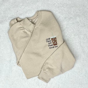 Könnte beinhalten: Ein beiger Pullover mit einer braunen und weißen Illustration eines Glases Eiskaffees und dem Text "Ooh They Got That Good Ice", der auf der Vorderseite gestickt ist.