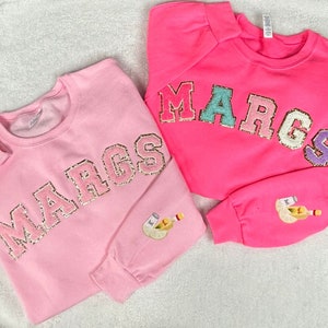 May include: Two pink sweatshirts with the word "MARGS" in sparkly letters. One sweatshirt has the letters in gold and the other has the letters in different colors. Both sweatshirts have a small embroidered image of a margarita.