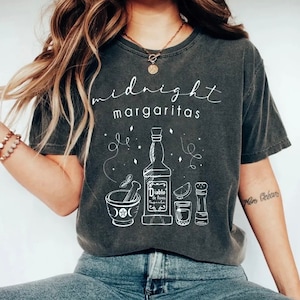 Puede incluir: Una camiseta gris oscuro con el texto "midnight margaritas" impreso en blanco. La camiseta presenta una ilustración blanca de una botella de tequila, una lima, un salero, un vaso de chupito y un mortero y una maja.