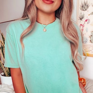 K&ouml;nnte beinhalten: Eine Frau tr&auml;gt ein hellblaues, kurz&auml;rmeliges T-Shirt mit einer goldenen Kettenkette und einem runden Anh&auml;nger.