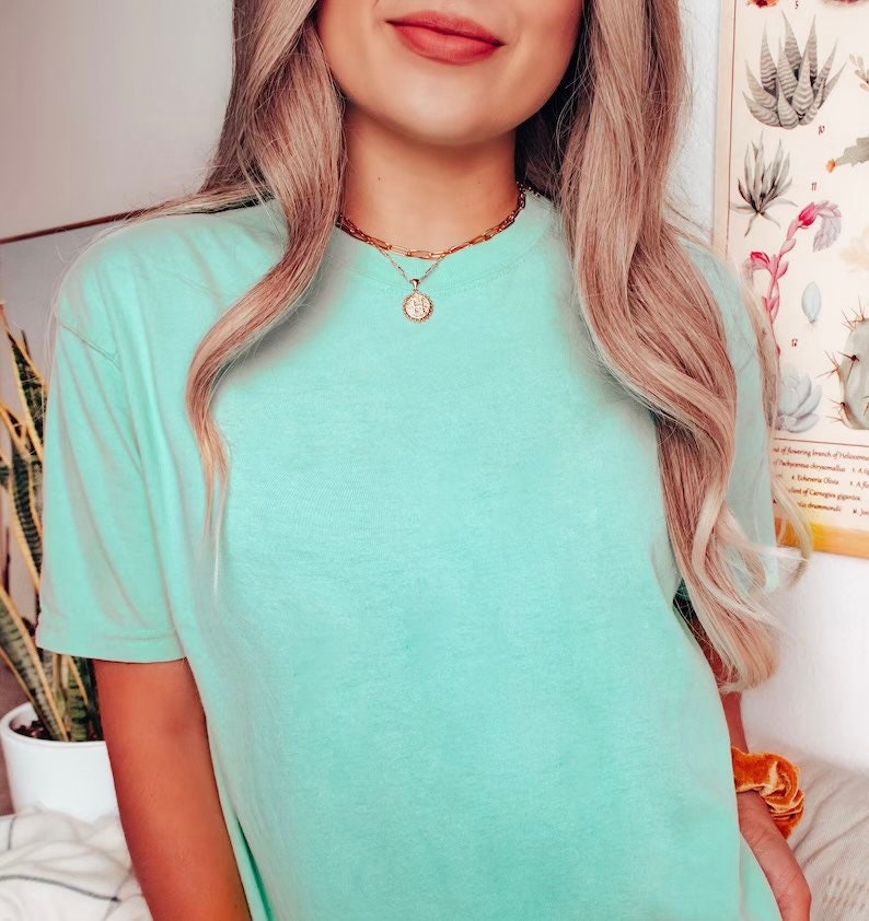 K&ouml;nnte beinhalten: Eine Frau tr&auml;gt ein hellblaues, kurz&auml;rmeliges T-Shirt mit einer goldenen Kettenkette und einem runden Anh&auml;nger.
