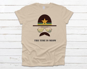 May include: A tan t-shirt with a graphic of a cartoon ranger wearing a brown hat with a gold star and green stripe, sunglasses, and a mustache. The text "THE TIME IS MEOW" is printed below the graphic.
