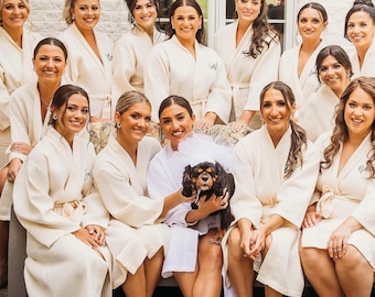 May include: A group of women wearing matching cream-colored robes, some with personalized initials embroidered on the front. They are gathered outdoors, with a small dog in the center. The setting appears to be a pre-wedding gathering.