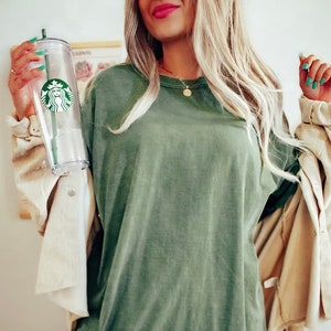 K&ouml;nnte beinhalten: Eine Frau tr&auml;gt ein gr&uuml;nes Langarmshirt und eine beige Cordjacke. Sie h&auml;lt einen durchsichtigen wiederverwendbaren Becher mit einem gr&uuml;nen Starbucks-Logo.