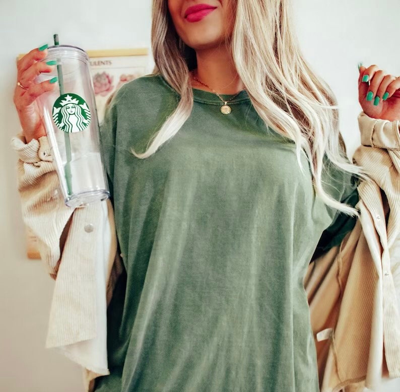 K&ouml;nnte beinhalten: Eine Frau tr&auml;gt ein gr&uuml;nes Langarmshirt und eine beige Cordjacke. Sie h&auml;lt einen durchsichtigen wiederverwendbaren Becher mit einem gr&uuml;nen Starbucks-Logo.