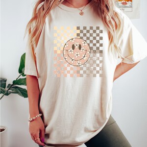 May include: A cream coloured t-shirt with a checkered pattern and a pink smiley face with white flowers. The smiley face is on a faded pink background.