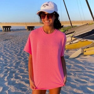 K&ouml;nnte beinhalten: Eine Frau tr&auml;gt ein pinkfarbenes T-Shirt und eine wei&szlig;e Baseballkappe mit der Aufschrift "NEW YORK NY". Sie steht an einem Sandstrand mit Segelbooten im Hintergrund.