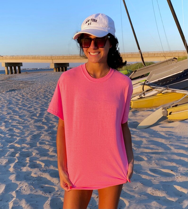 K&ouml;nnte beinhalten: Eine Frau tr&auml;gt ein pinkfarbenes T-Shirt und eine wei&szlig;e Baseballkappe mit der Aufschrift "NEW YORK NY". Sie steht an einem Sandstrand mit Segelbooten im Hintergrund.
