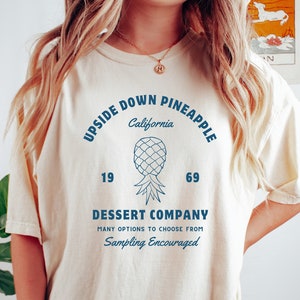 Op de afbeelding: Een beige T-shirt met een blauw grafisch ontwerp met de tekst "Upside Down Pineapple California" met een ananasillustratie en de nummers 19 en 69. De tekst onder de ananas luidt "Dessert Company Many Options to Choose From Sampling Encouraged".