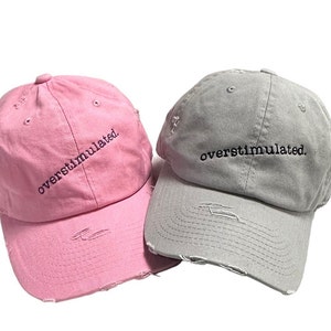 May include: Two distressed baseball caps, one pink and one gray, with the text "overstimulated." embroidered on the front.