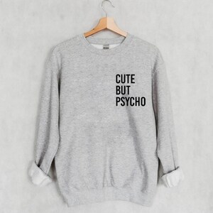 K&ouml;nnte beinhalten: Grauer Pullover mit dem Aufdruck "CUTE BUT PSYCHO" in Schwarz auf der Vorderseite.