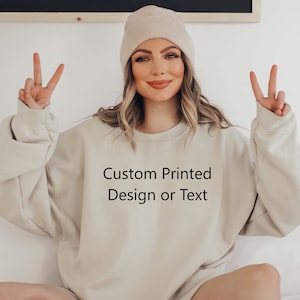 以下が含まれることがあります： 「Custom Printed Design or Text」の文字が入ったベージュのスウェットシャツ。スウェットシャツは長袖でクルーネックです。画像に写っている人はベージュのビーニー帽をかぶっています。