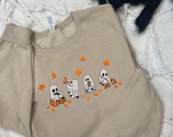 May include: A tan sweatshirt featuring a design of five ghosts dressed in autumnal attire. The ghosts are holding pumpkins, leaves, and a cup of coffee. The sweatshirt has long sleeves and a crew neck.