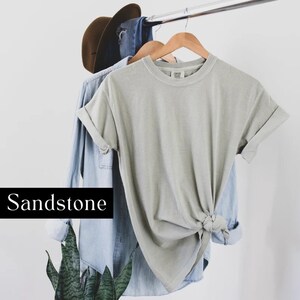 K&ouml;nnte beinhalten: Ein hellgraues T-Shirt mit hochgekrempelten &Auml;rmeln, das an einem Kleiderb&uuml;gel h&auml;ngt. Das T-Shirt ist unten verknotet und hat den Text "COMFORT COLORS" auf dem Etikett. Der Text "Sandstone" ist unter dem T-Shirt gedruckt.