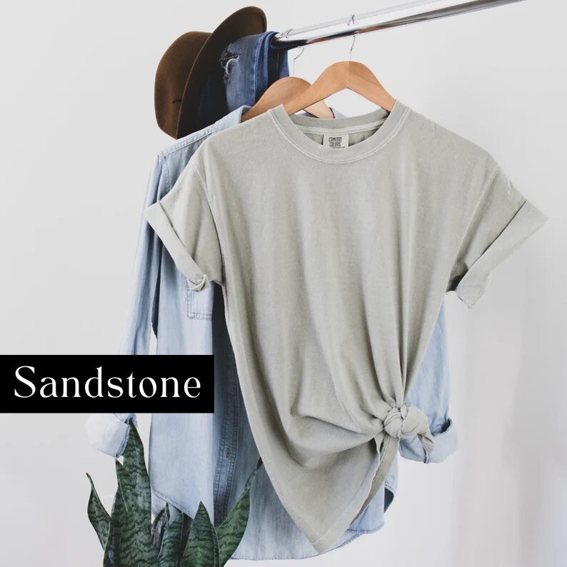 K&ouml;nnte beinhalten: Ein hellgraues T-Shirt mit hochgekrempelten &Auml;rmeln, das an einem Kleiderb&uuml;gel h&auml;ngt. Das T-Shirt ist unten verknotet und hat den Text "COMFORT COLORS" auf dem Etikett. Der Text "Sandstone" ist unter dem T-Shirt gedruckt.