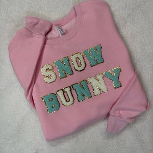 Peut inclure: Sweat-shirt rose avec le texte "SNOW BUNNY" en lettres bleues et dorées pailletées.