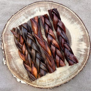 May include: Five braided dog chews in varying shades of brown and reddish-brown are arranged on a rustic wooden slice. The chews have a textured, woven appearance, and the wood slice has a natural, unfinished edge.