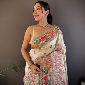 Pode incluir: Sari de cor marfim com uma borda detalhada com motivos florais e animais coloridos. O sari tem um design bordado com tons de rosa, verde, laranja e azul. O tecido drapeia elegantemente, mostrando o artesanato intrincado.