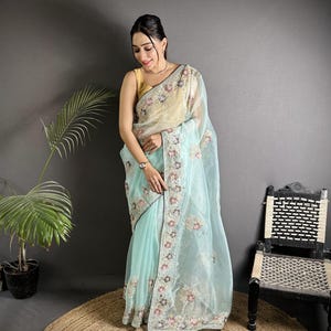 May include: A light blue saree with intricate floral embroidery along the borders and a sheer overlay. The saree features a gold-colored blouse and is draped elegantly. The design includes a detailed floral pattern with pink, white, and gold accents.